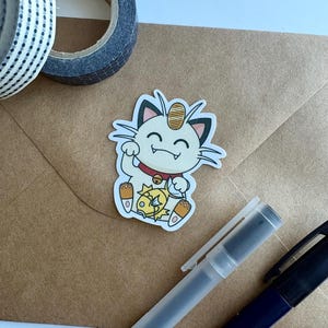 Puede incluir: Una pegatina troquelada con un gato de dibujos animados con una moneda de oro en la cabeza, un collar rojo y una pequeña criatura en sus brazos. La pegatina está sobre un sobre marrón, con cinta washi y bolígrafos en el fondo.
