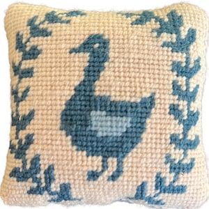 May include: A small, square, needlepoint pillow featuring a blue goose design on a light pink background. The goose is surrounded by a border of blue leaves. The pillow has a textured appearance.