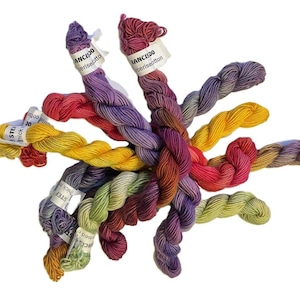 Pode incluir: Uma coleção de novelos de fios coloridos em vários tons de vermelho, amarelo, roxo e verde. Cada novelo tem uma etiqueta com texto. O fio é disposto em um padrão circular sobre um fundo branco, mostrando as cores vibrantes.