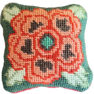 May include: A square, handmade needlepoint pillow featuring a large, stylized flower. The flower is primarily coral and peach, with dark brown and teal accents. The background is green with light green leaves, and the edges are a darker green.