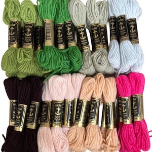 Op de afbeelding: Een verzameling kleurrijke Anchor Tapisserie Wool-Laine borduurgaren strengen. De strengen zijn in verschillende tinten groen, bruin, roze, perzik en wit. Elke streng is voorzien van het merk Anchor en een productnummer.