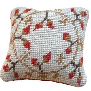 May include: A small, square pillow with a white background and a floral design. The design features brown branches with red flower clusters. The pillow has a red border and is made of a textured fabric.