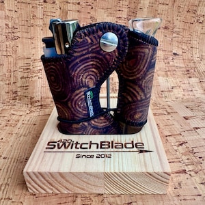 May include: A brown cork-textured holder with a dark circular pattern, featuring a silver metal screw, a lighter, and a glass piece. The holder sits on a light-colored wooden base with the laser-engraved text 420 SwitchBlade Since 2012.