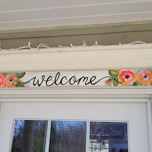 May include: A wooden welcome sign with a white background and blue and white stripes. The sign is decorated with painted flowers in shades of orange, pink, and yellow. The word "welcome" is written in black cursive lettering.