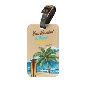 Op de afbeelding: Beige bagagelabel met een zwarte riem en een gouden gesp. De label toont een tropische scène met een palmboom, surfboard en golven. De tekst luidt "Even the wind & Waves Obey Him - Mark 4:41."
