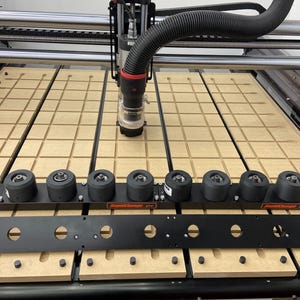 Könnte beinhalten: Eine CNC-Maschine mit einem beigefarbenen Rastertisch und einem schwarzen und silbernen Portal. Ein schwarzer Schlauch ist mit der Spindel verbunden. Ein schwarzer Metallstab mit kreisförmigen Aussparungen und eine Reihe schwarzer Räder befinden sich im Vordergrund.