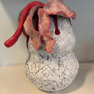 May include: A unique ceramic sculpture with a textured white surface and a cracked pattern. The sculpture features red and pink organic shapes and drips. The artwork is a decorative piece, possibly for a home or gallery.