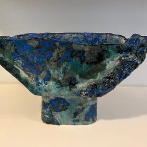May include: A textured, handmade ceramic bowl with a wide, open top and a pedestal base. The bowl features a blend of teal, blue, and black colors, with a rough, uneven surface. The design suggests a unique, artistic piece for home decor.