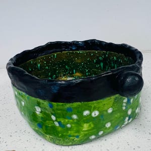 Peut inclure: Un bol en céramique fait à la main avec un bord noir et un extérieur vert. Le bol présente des accents mouchetés blancs et bleus sur la surface verte. Une petite poignée noire ronde est sur le côté. L'intérieur est vert foncé.