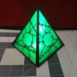 May include: A green, pyramid-shaped decorative object with a black outline. The pyramid has a barbed wire design on each face. The object is illuminated with a bright green light, creating a striking visual effect. The background is a red surface.