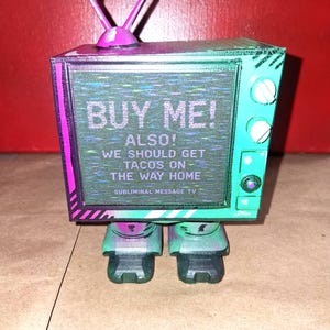 May include: A teal and purple television-shaped figure with two antennas on top. The screen displays the text: BUY ME! ALSO! WE SHOULD GET TACOS ON THE WAY HOME SUBLIMINAL MESSAGE TV. The figure has two knobs and a small circular button on the side.