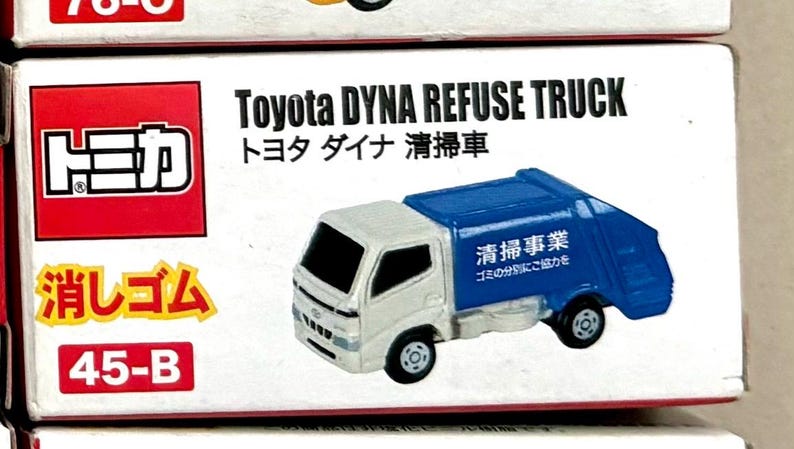 May include: A miniature Toyota Dyna Refuse Truck toy. The truck has a white cab and a blue refuse container. Text on the box includes "Toyota DYNA REFUSE TRUCK" and Japanese characters. The box also has the number "45-B".