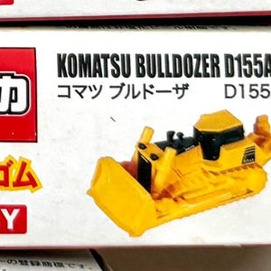 May include: A yellow Komatsu Bulldozer D155AX-6 toy model. The toy is on a white box with red and yellow text. The box also has Japanese text. The bulldozer has a black cab and a blade.