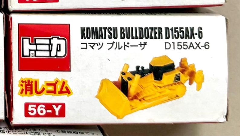 May include: A yellow Komatsu Bulldozer D155AX-6 toy model. The toy is on a white box with red and yellow text. The box also has Japanese text. The bulldozer has a black cab and a blade.