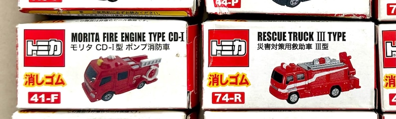 May include: Close-up of two toy car boxes. The left box features a red fire engine with the text "MORITA FIRE ENGINE TYPE CD-I". The right box shows a red rescue truck with the text "RESCUE TRUCK III TYPE". Both boxes have Japanese text.