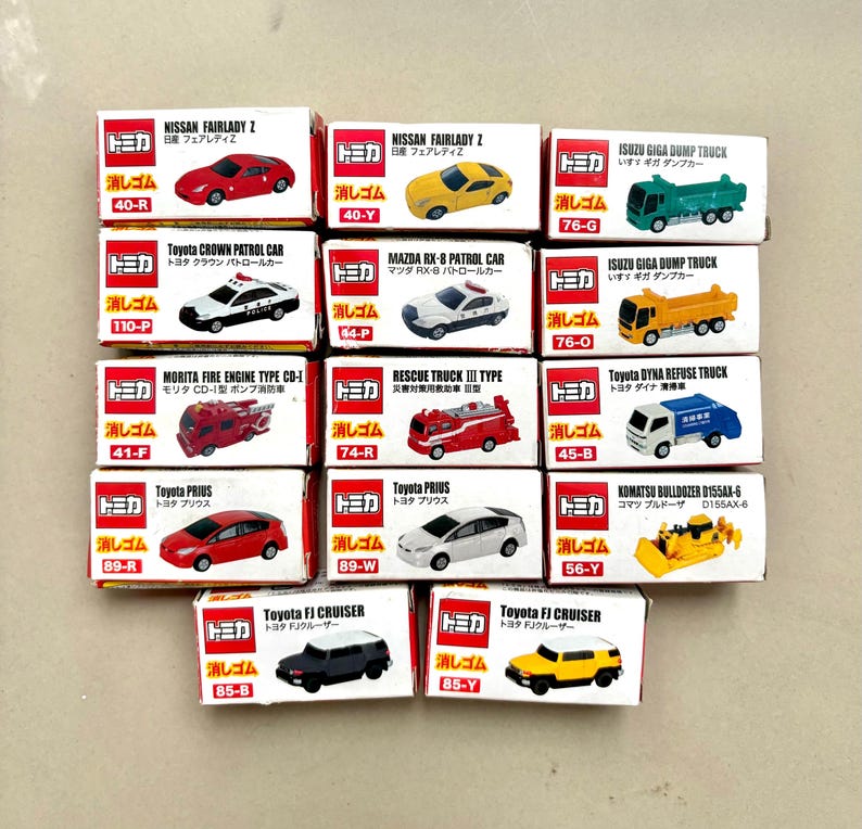 May include: A collection of miniature die-cast toy cars in their original boxes. The cars include various models like Nissan Fairlady Z, Toyota Prius, and Isuzu Giga Dump Truck. The boxes are white with Japanese text and illustrations of the cars.