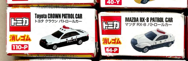 May include: Two toy car boxes. One box features a white and black Toyota Crown Patrol Car with the word "POLICE" on the side. The other box shows a white and black Mazda RX-8 Patrol Car.