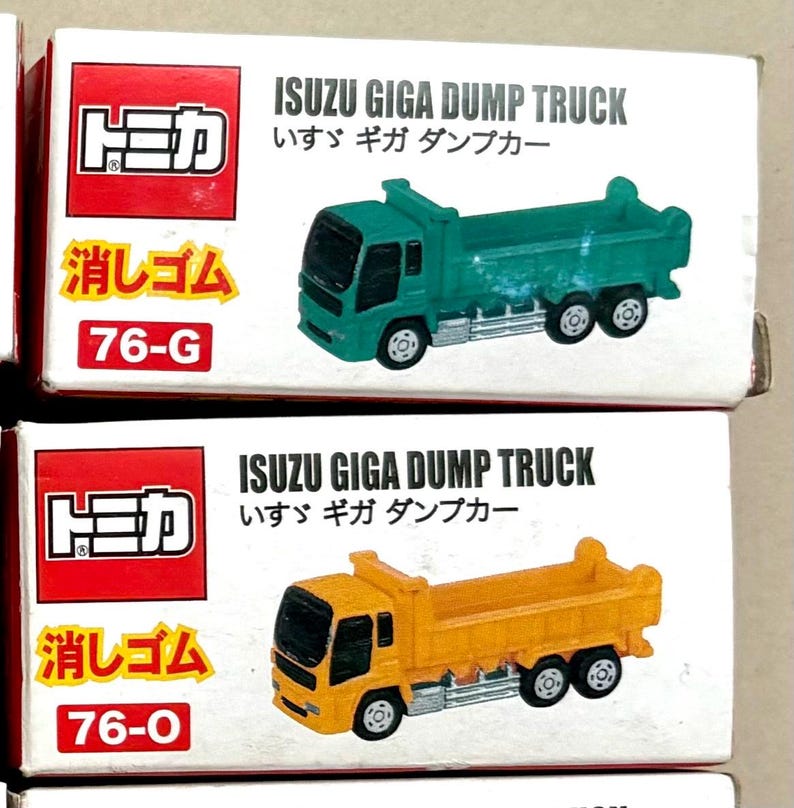 May include: Two toy ISUZU GIGA DUMP TRUCK models in their original boxes. One truck is green, the other is yellow. The boxes have Japanese text and the number 76-G and 76-O. The boxes are red and white.
