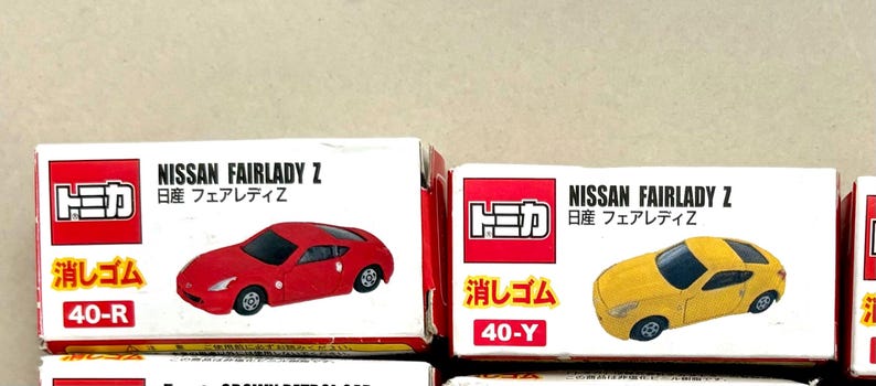 May include: Boxes of toy cars, featuring a red and a yellow Nissan Fairlady Z model. Each box has the car's image, the brand name, and Japanese text. The boxes are white with red accents.
