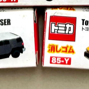 May include: Two toy car boxes. One box features a black Toyota FJ Cruiser model with a white roof. The other box features a yellow Toyota FJ Cruiser model with a white roof. The boxes have Japanese text and the text "Toyota FJ CRUISER".