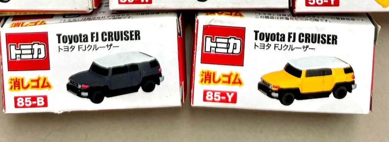May include: Two toy car boxes. One box features a black Toyota FJ Cruiser model with a white roof. The other box features a yellow Toyota FJ Cruiser model with a white roof. The boxes have Japanese text and the text "Toyota FJ CRUISER".