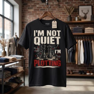 May include: A black short-sleeved t-shirt featuring a graphic print with the text I'M NOT QUIET I'M PLOTTING. The design includes two chess pieces on a board, tic-tac-toe grids, and red and white accents. A Boston Clothiers tag is attached to the collar.