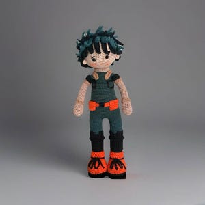May include: A handmade crochet doll of a character with green hair and a green jumpsuit with orange accents. The doll has black boots with orange details. The doll is standing against a gray background.