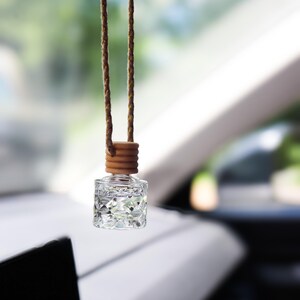 Puede incluir: Un frasco pequeño de vidrio transparente con una textura facetada similar a un diamante, colgado de un cordón trenzado marrón. El frasco tiene una tapa de madera clara y está suspendido del espejo retrovisor de un vehículo con líquido.