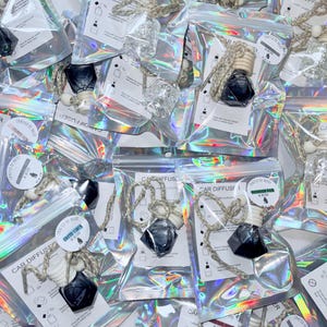 Może przedstawiać: Zbliżenie na kilka małych, przezroczystych szklanych butelek z czarnymi zakrętkami i przymocowanym białym sznurkiem. Butelki są wypełnione przezroczystym płynem i zapakowane w indywidualne, przezroczyste plastikowe torebki z holograficznym wykończeniem. Torebki są oznaczone tekstem "CAR DIFFUSER" i "CRAFTED BY AESTHETICS".