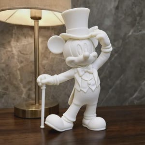 Puede incluir: Una figura de color blanco mate de un personaje de ratón de dibujos animados que lleva un esmoquin formal, una pajarita y un sombrero de copa. El personaje está de pie con una mano tocando el ala del sombrero y la otra sobre un bastón fino.