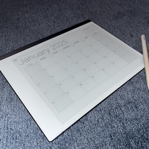 Puede incluir: Un calendario digital blanco con un borde negro y el texto "Enero 2025" en la parte superior. Un lápiz blanco está acostado junto al calendario sobre una superficie azul.