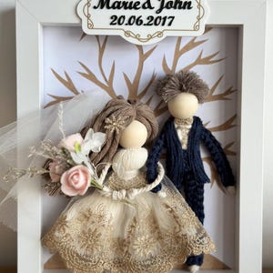 Puede incluir: Un marco blanco con un árbol marrón y dos muñecas de hilo vestidas de novia y novio. La novia lleva un vestido blanco y dorado y el novio lleva un traje azul marino. El marco tiene un cartel que dice "Marie & John 20.06.2017".