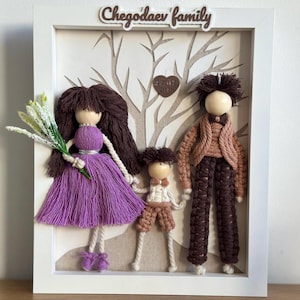 May include: A framed artwork featuring a family of three yarn dolls. The dolls are dressed in purple, brown, and beige. The background is a beige fabric with a tree design. The frame is white and has the text "Chegoddaev family" at the top.