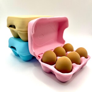 May include: Three colorful egg cartons, stacked. A pink carton holds six brown eggs. A blue carton and a yellow carton are stacked below. The cartons are made of a textured material.