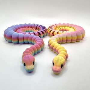 May include: Two colorful, articulated snake toys in rainbow hues. Each snake is coiled and has a detailed head with black eyes. The toys are made of layered, textured material, creating a 3D effect. The colors transition smoothly from pink to yellow and blue.