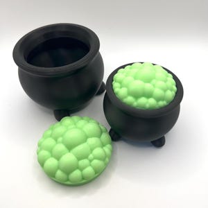 May include: Three-piece set of black cauldron-shaped containers. One cauldron is empty, another is filled with a light green, bubbly substance, and the third is a lid with a matching bubbly texture. The set is likely for Halloween or themed decor.