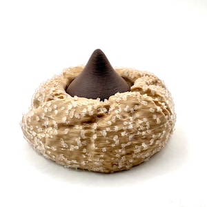 May include: A brown, cone-shaped object rises from the center of a beige, textured, circular form. The circular form is adorned with small, clear, crystalline embellishments, creating a unique, decorative piece. The background is plain white.