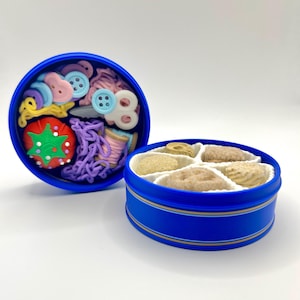 May include: Two blue tins, one open and one closed, on a white surface. The open tin contains sewing notions like buttons, thread, and a tomato pin. The closed tin holds several cookies. The tins have a blue and gold stripe design.