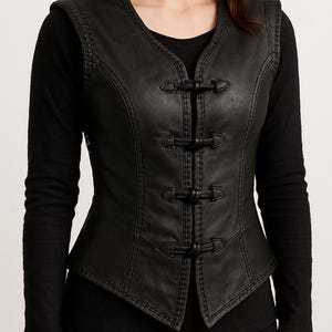 Puede incluir: Chaleco de cuero negro con cuello en V y tres cierres de palanca. El chaleco tiene una silueta ajustada con costuras decorativas a lo largo de las costuras. El chaleco se usa sobre una camisa negra de manga larga.