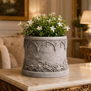 May include: A cylindrical grey stone planter featuring an embossed relief of climbing rose vines and gothic-style arches along the top rim. The planter is filled with a lush green plant displaying numerous small, white, five-petaled flowers.