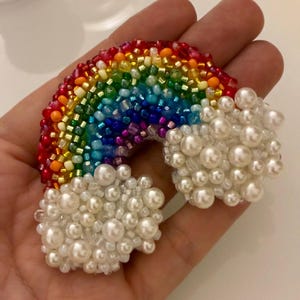 Può includere: Una spilla arcobaleno artigianale con perline di vetro rosse, arancioni, gialle, verdi, blu e viola disposte ad arco, affiancata da due nuvole soffici realizzate con perle bianche artificiali e perline di vetro trasparenti.