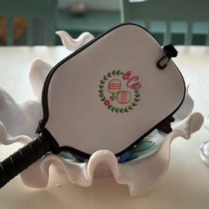 Pode incluir: Estojo branco para raquete de pickleball com zíper e alça pretos. O estojo apresenta uma coroa verde com duas raquetes de pickleball e as iniciais "BCR" bordadas em rosa.