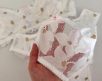 May include: White lace lingerie set with floral and heart patterns. The bra has pink straps and bows, and the matching bottoms are held by a hand. The set features gold heart accents and a delicate, feminine design.