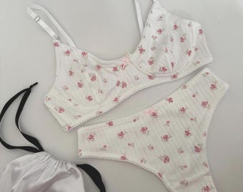 May include: A white bra and thong set with a delicate floral pattern in pink. The set includes a matching drawstring bag. The bra has adjustable straps and a small bow. The thong has a scalloped edge.