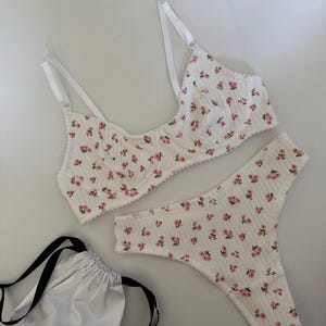 May include: White floral-print lingerie set featuring a bra and thong. The bra has adjustable straps and underwire support. The set is adorned with small pink flowers. A white satin drawstring bag with black straps is also included.