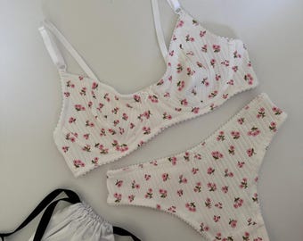 May include: White floral-print lingerie set featuring a bra and thong. The bra has adjustable straps and underwire support. The set is adorned with small pink flowers. A white satin drawstring bag with black straps is also included.