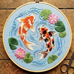 May include: Embroidered artwork of two koi carp in a tranquil aquatic scene. The fish are orange, white, and black, amidst blue water, green lily pads, and pink lotus flowers. The embroidery is set in a wooden hoop.