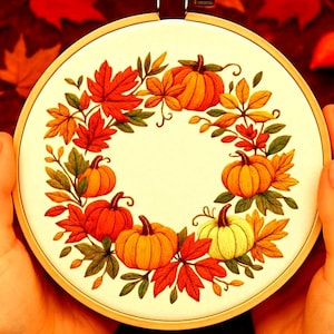 Könnte beinhalten: Ein handgestickter Stickrahmen mit einem herbstlichen Design. Das Design zeigt orangefarbene Kürbisse und bunte Herbstblätter in Rot-, Orange- und Gelbtönen, die in einem kreisförmigen Kranz auf cremefarbenem Hintergrund angeordnet sind.