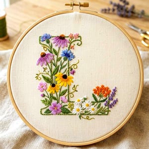May include: Embroidered floral letter "L" in a wooden hoop. The letter is filled with colorful flowers, including purple coneflowers, yellow black-eyed susans, and daisies. The embroidery is on a cream-colored fabric.