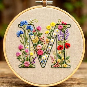 Op de afbeelding: Geborduurde bloemenletter "M" in een houten hoepel. De letter is gevuld met kleurrijke bloemen, waaronder blauwe korenbloemen, roze rozen en rode klaprozen. De achtergrond is een neutrale beige stof.